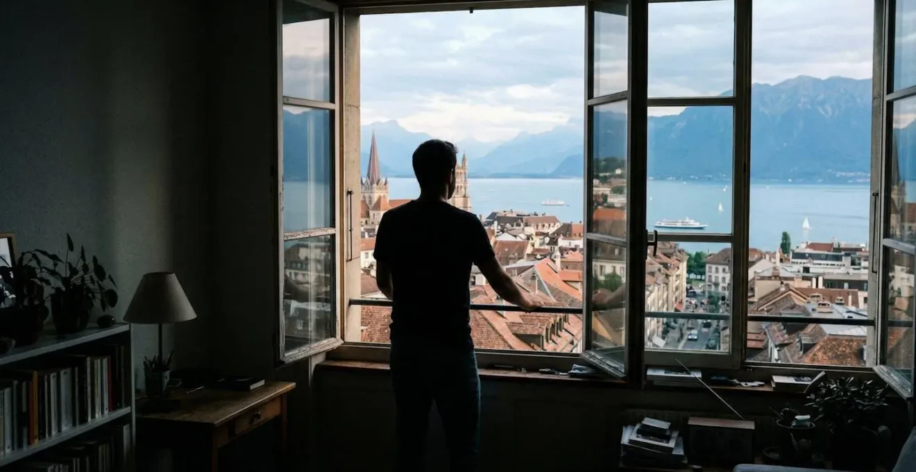 Une personne de dos regarde par la fenêtre d'un appartement avec vue sur les toits d'une ville suisse et le lac en arrière-plan